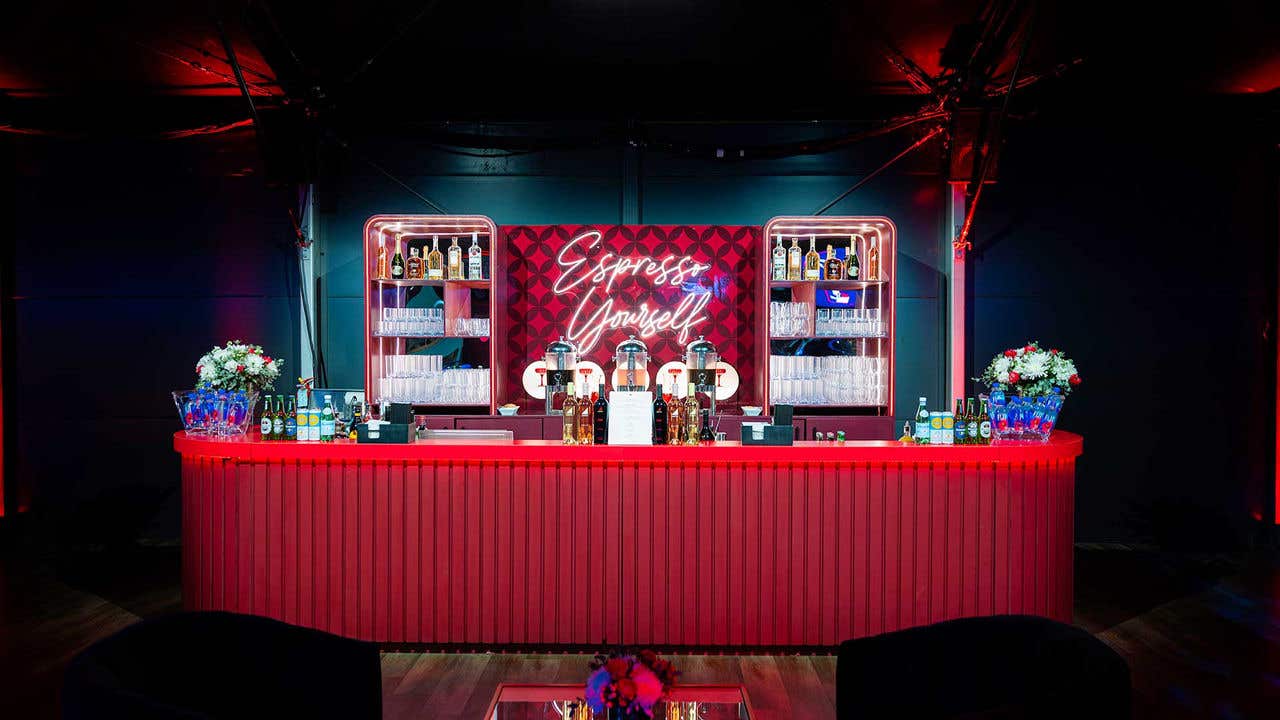 A red bar with an assortment of drinks, with a neon sign in the back saying "Espresso Yourself"