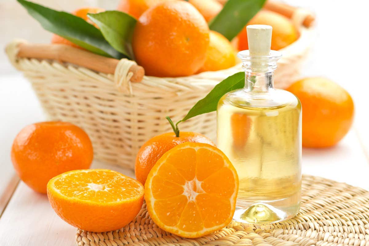 A bottle of citrus oil next to fresh oranges, some whole and one halved, on a woven mat with a basket of oranges in the background.