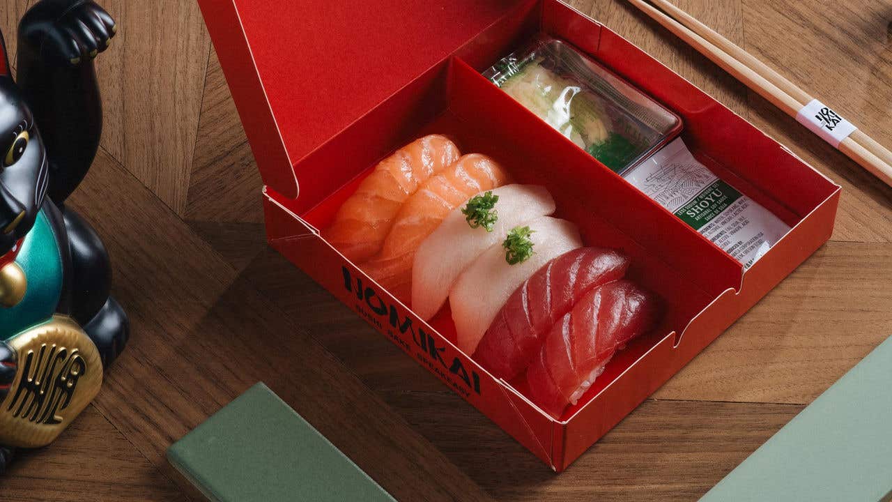 An open red box containing assorted sushi on a wooden table, with a Maneki-neko figurine and chopsticks nearby.