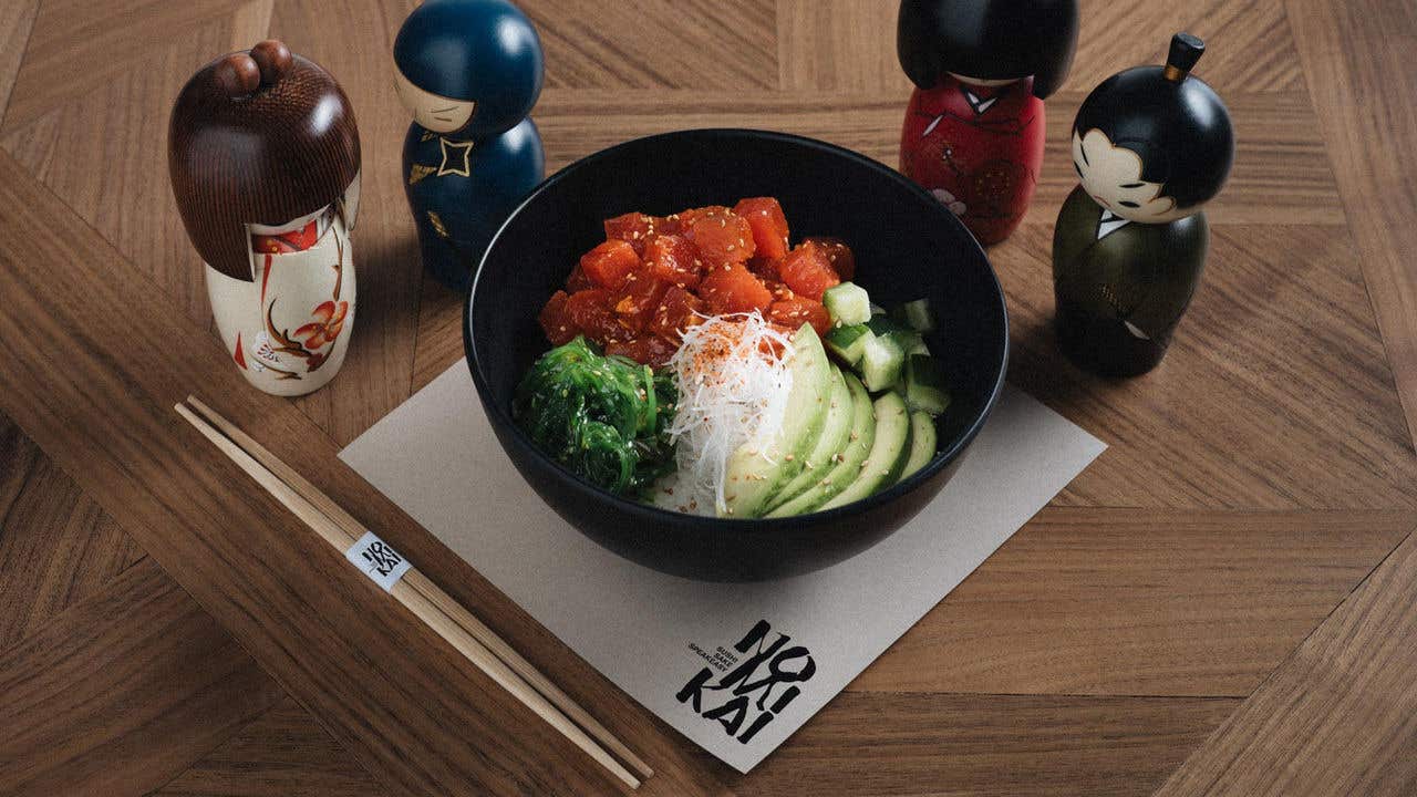 Black bowl with tuna, avocado, and seaweed surrounded by wooden Japanese figurines on a patterned wood table.