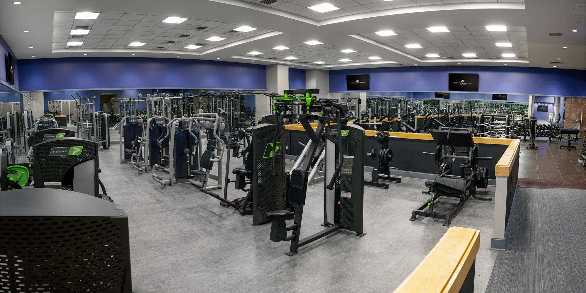 Panoramic view of a modern, spacious gym featuring various strength training machines and weightlifting equipment, with mirrored walls and flat-screen TVs mounted throughout.