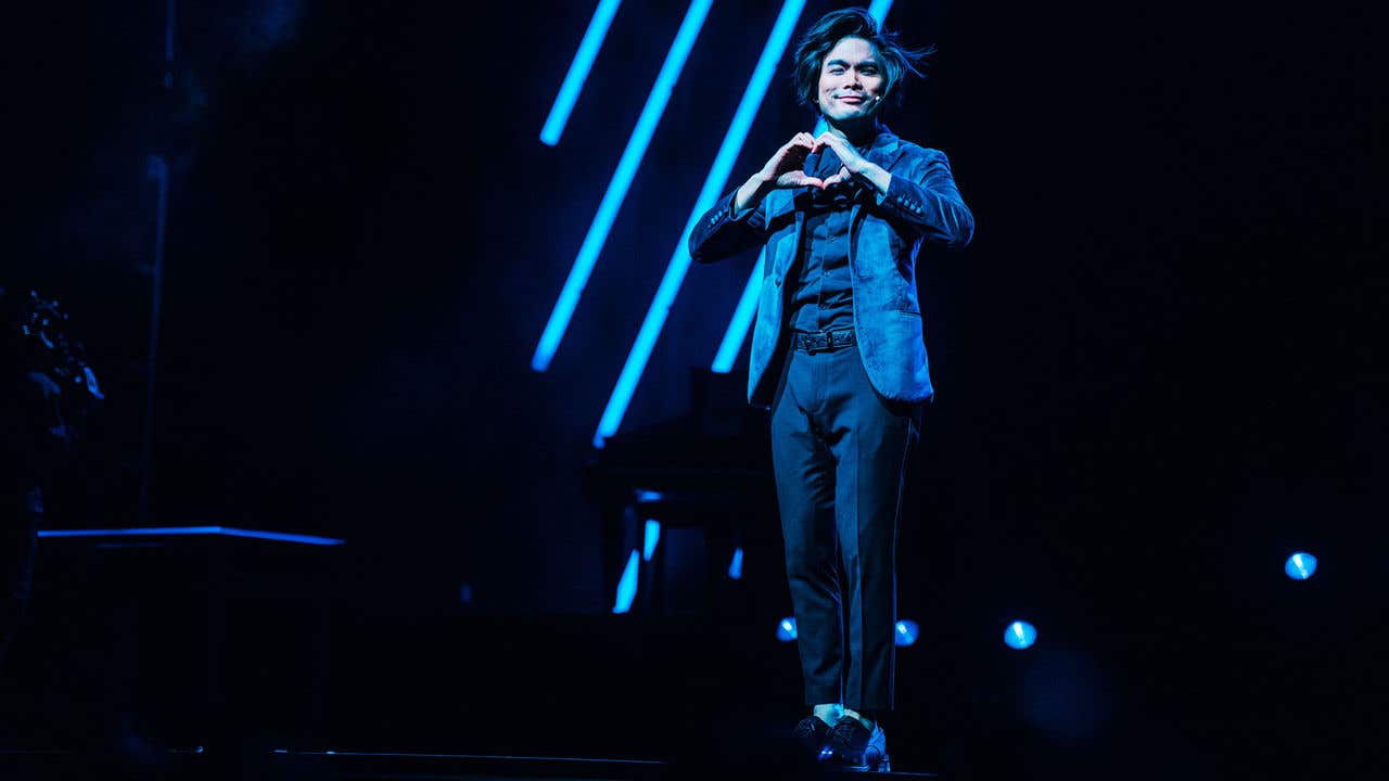 Shin Lim dressed in all black wearing a dark blue jacket looking out at the audience and forming the shape of a heart with his hands. 