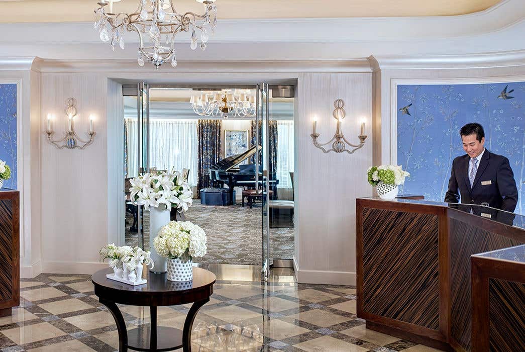 Elegant las vegas hotel lobby with two wooden reception desks, floral arrangements, and a chandelier. A grand piano is in the background.