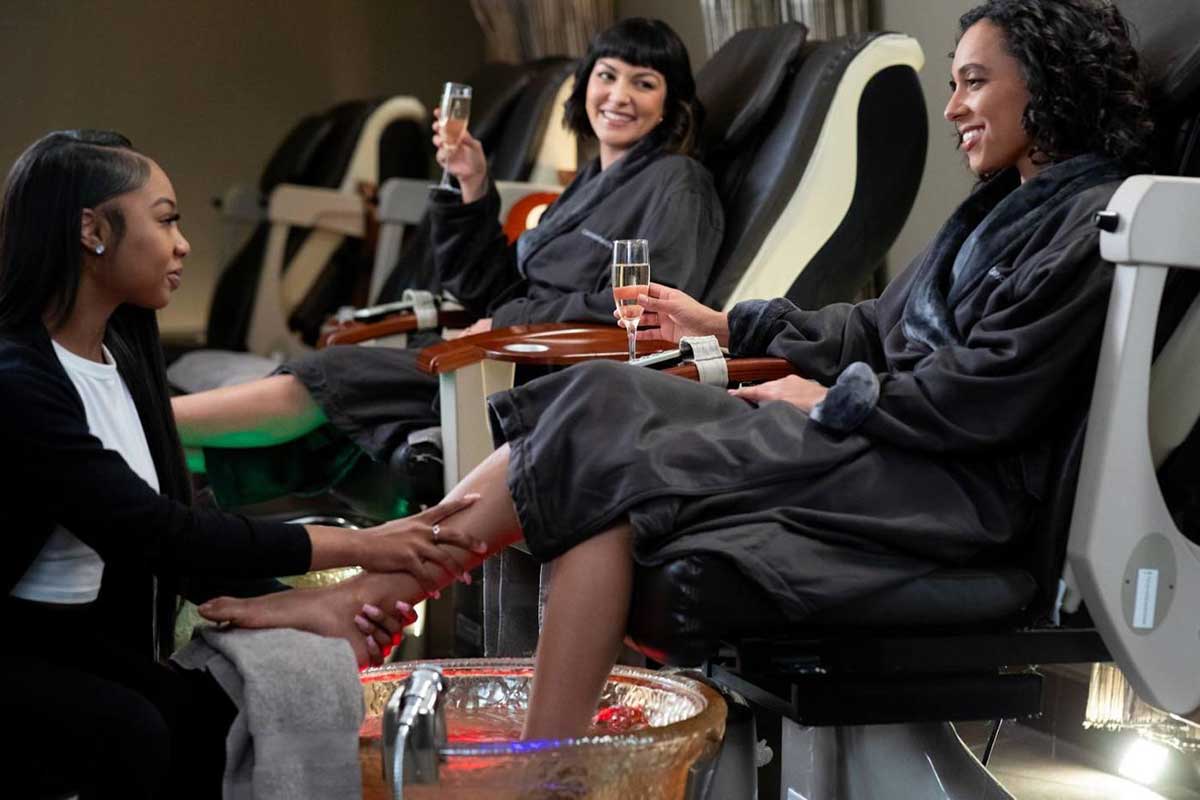 Two women in robes sit in nail salon chairs and receive a pedicure.