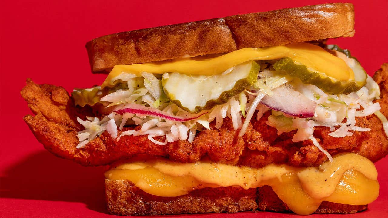 Large sandwich with fried chicken, pickles, cole slaw, cheese and a yellow sauce in front of a red background. 