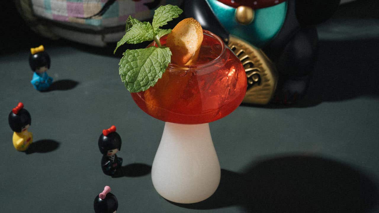A cocktail in a mushroom-shaped glass with mint and citrus peel garnish, surrounded by colorful decorative dolls.