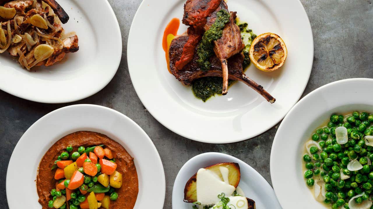 Five plated dishes including lamb chops, pork ribs, peas, root vegetables, and potatoes on a gray surface.