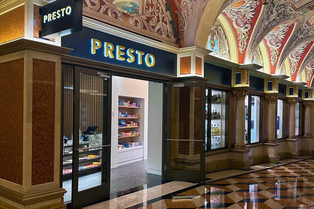 An open doorway to a shop named Presto, containing small convenient items, in a dim hallway lit from extremely decorative archways above