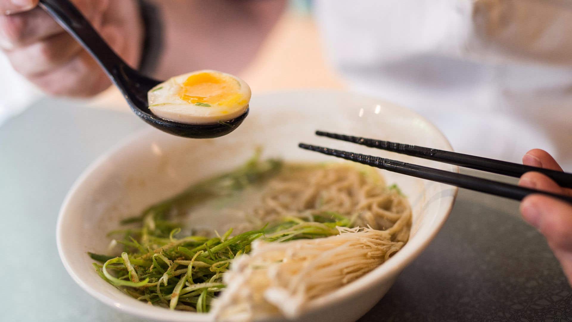 Ramen soup in a bowl with someone holding a hardboiled egg in a spoon and chopsticks over the bowl.