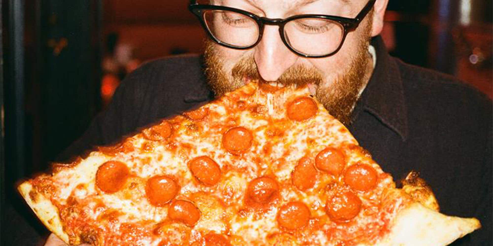 Man standing up biting into a large slice of pepperoni pizza.